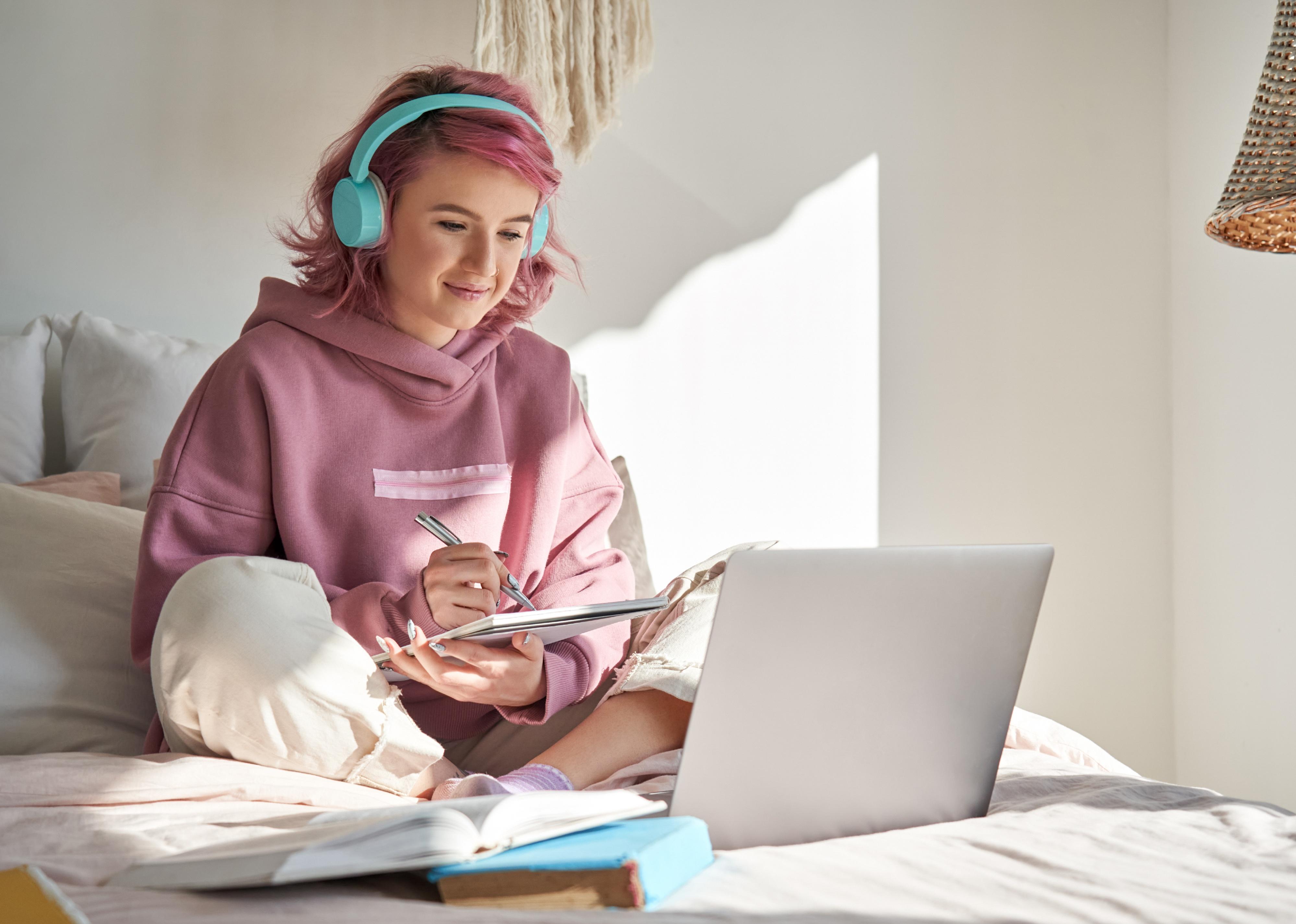 Student with pink hair with headphones takes notes while watching online webinar.