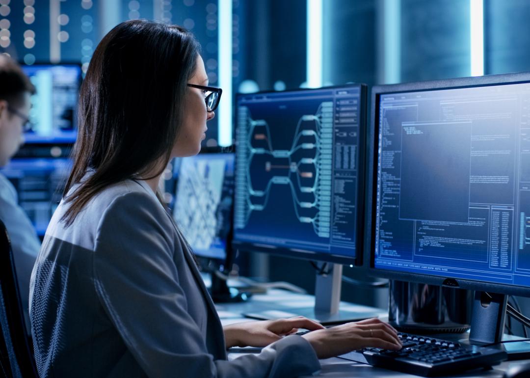 Over the shoulder angle of a business woman working in a monitoring room in front of computer screens.