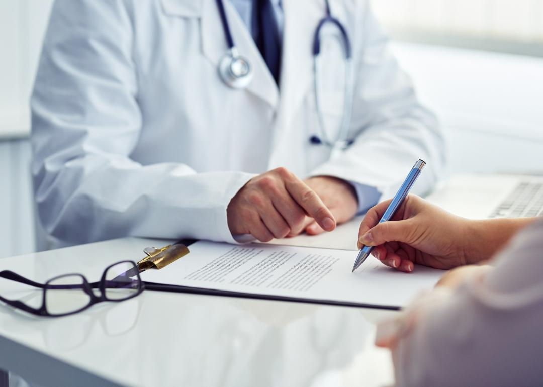 A patient in an office filling out medical document with doctor sitting across the table.