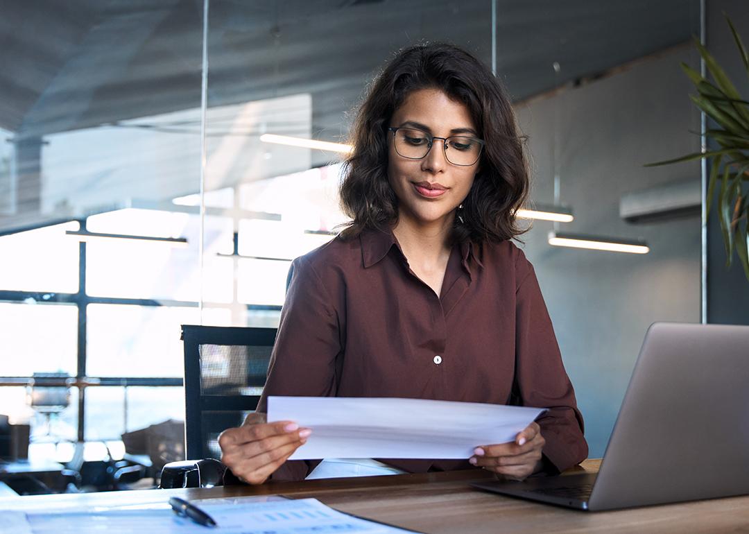 Focused young businesswoman reading financial report document and using a laptop in the workplace.