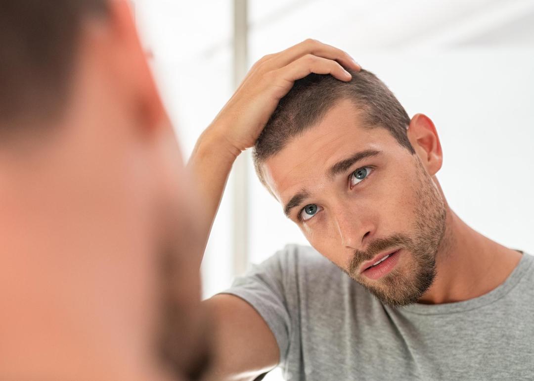 Young man with shaved head looking at mirror in bathroom at home, running fingers over hair..