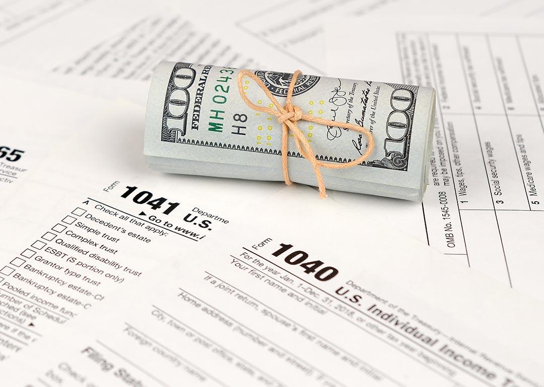A tied up roll of hundred dollar bills on top of various paper U.S. tax forms like the 1040 and 1041.