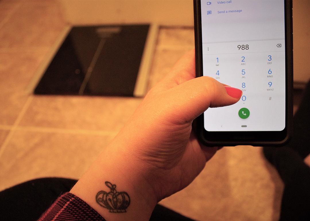 A woman with a tattoo dials the national suicide hotline '988' number from the floor in front of a body weight scale.