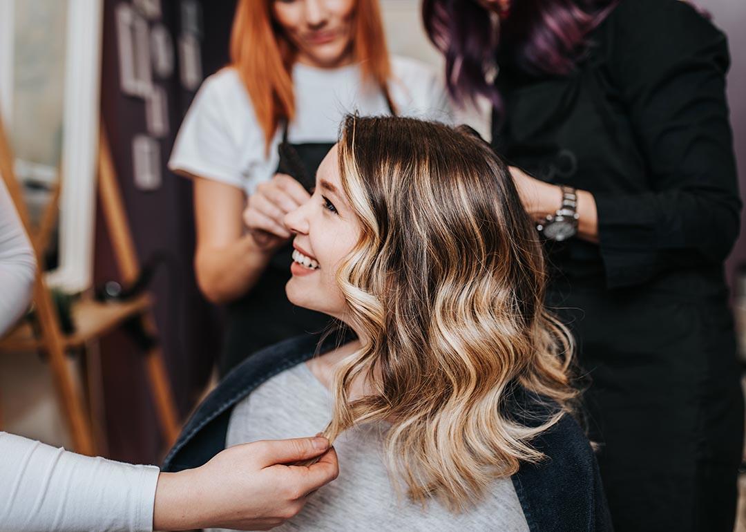 Side profile of woman with blond and brunette hair in long layers smiling at hair salon with two stylists behind her in the background.