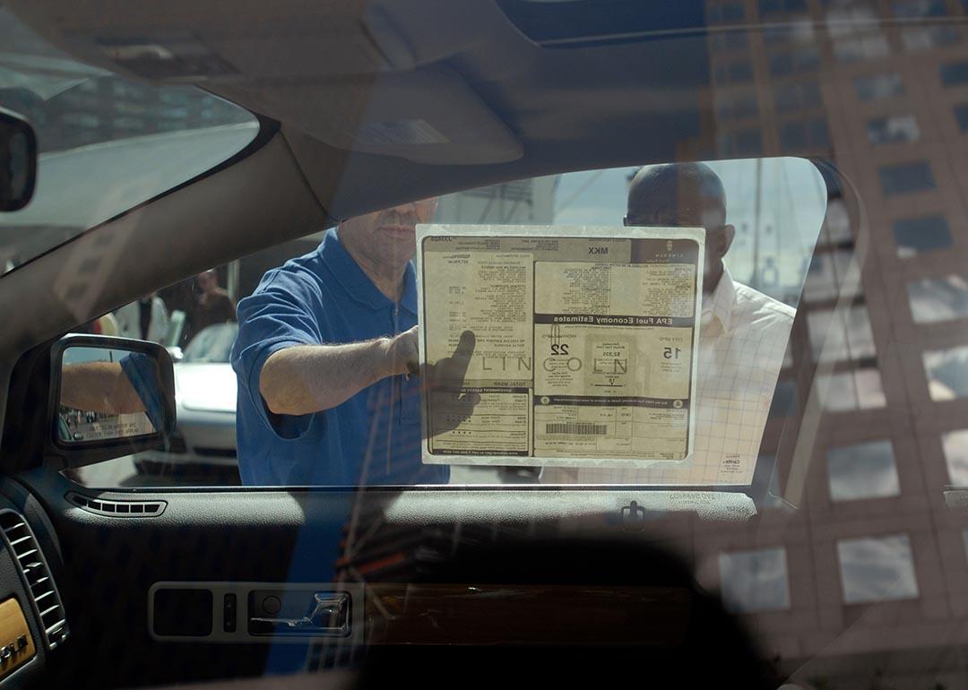 Interior view of car from driver's side looking out passenger window where two people are reading a sticker on the window with the car's features. 