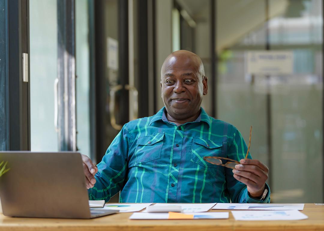 A senior man excitingly looks at financial documents while using a laptop.