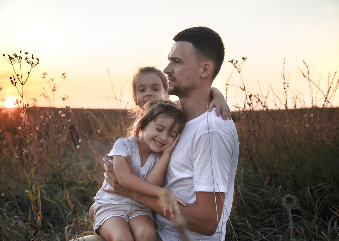 A father with her two young daughters out on a grass field during sunset.
