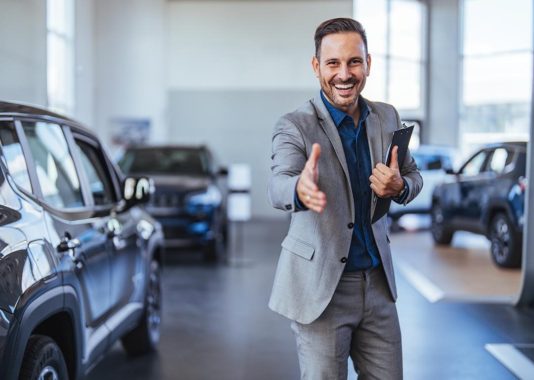 A friendly and cheerful salesman poses to greet car showroom clients with a handshake.