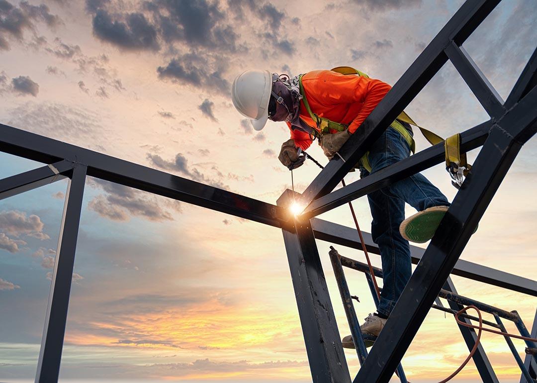 Low angle view of a welder is welding steel on a steel roof truss with the sunrise in the background.