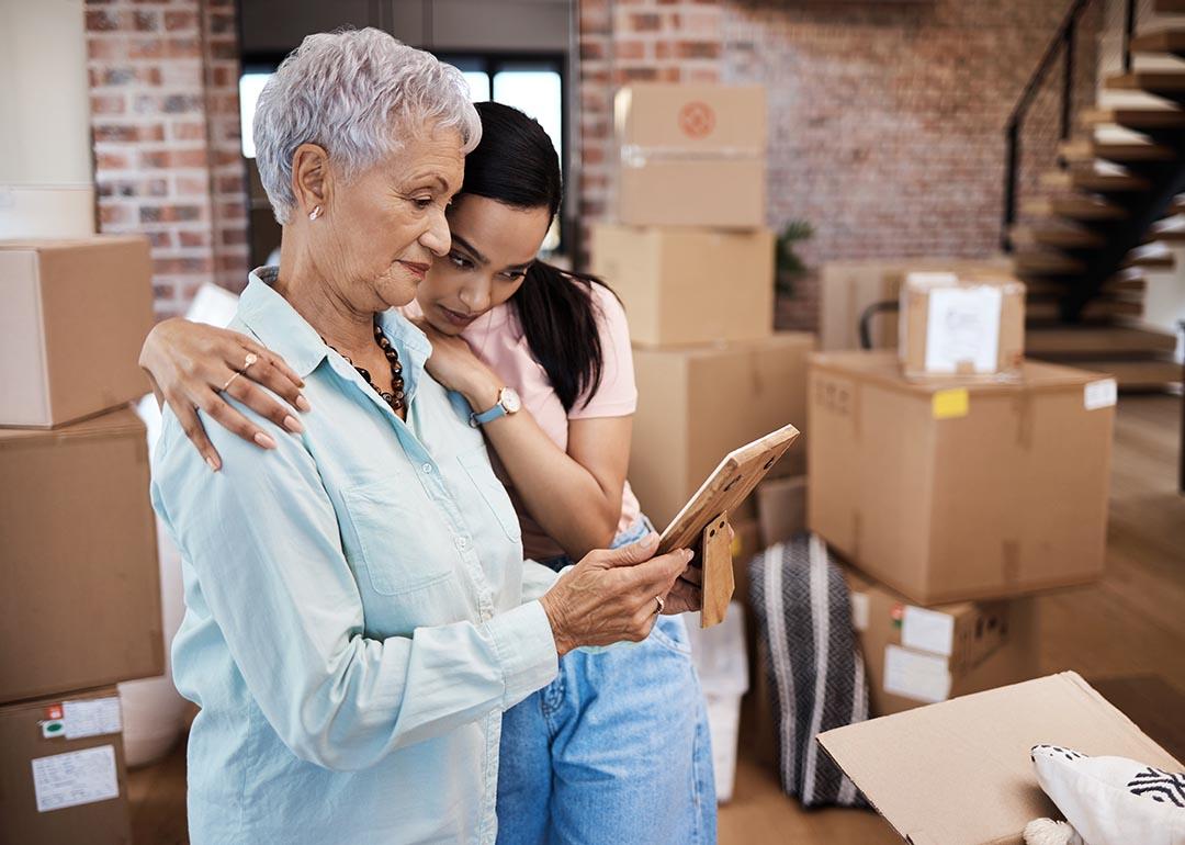 An older woman looking at a photograph while getting a hug from a younger family member while packing boxes on moving day.