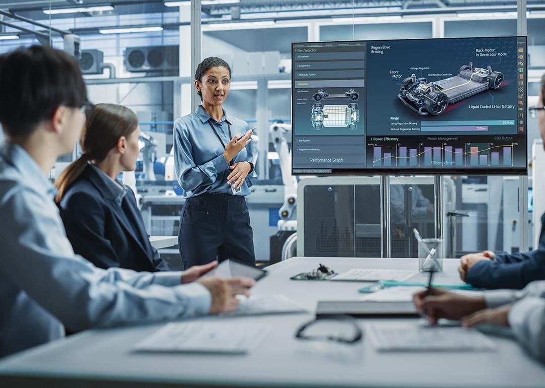 A female project manager leads her team of engineers with a presentation of a new car model in an EV manufacturing facility office.