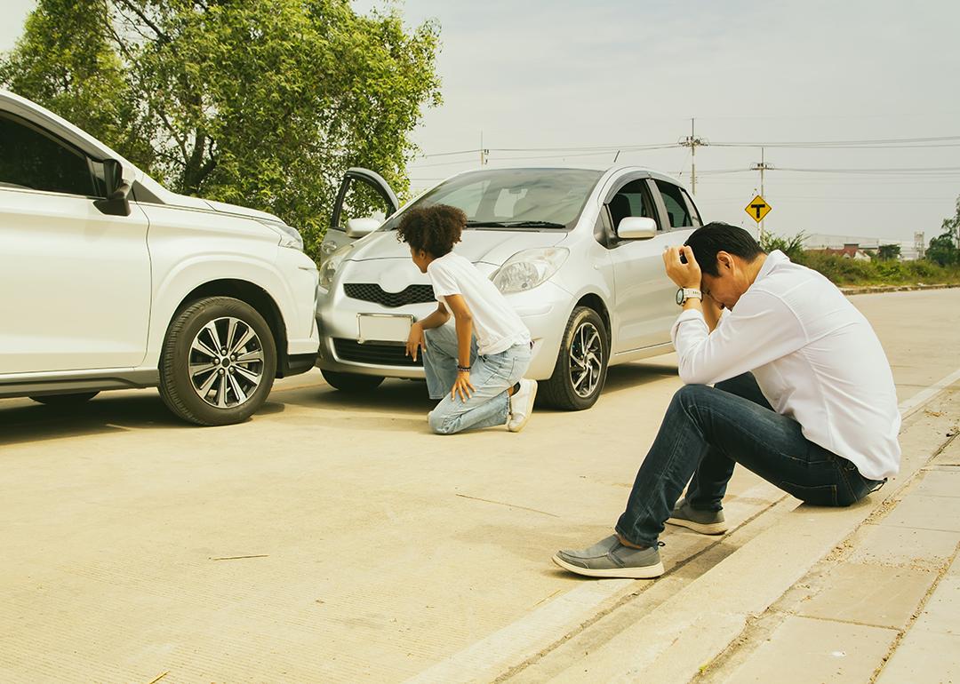 Drivers of two cars caught in a road accident inspect and sit down by a roadside in distress.