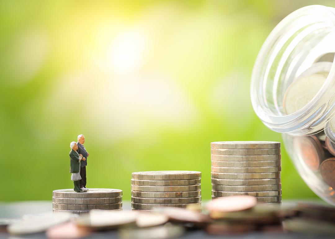 A miniature elderly couple stands on a stack of coins leading to a glass jar full of savings.
