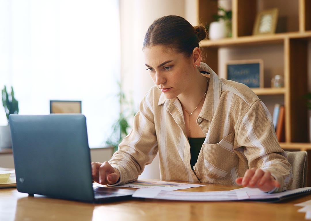 Determined-looking woman looking intently into laptop screen. She is sitting in an office with wooden bookshelves and a window behind her.