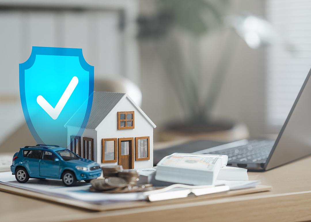 Cash, figures of a house and a car beside a laptop illustrated with a blue safety check graphic as a concept of insurance.