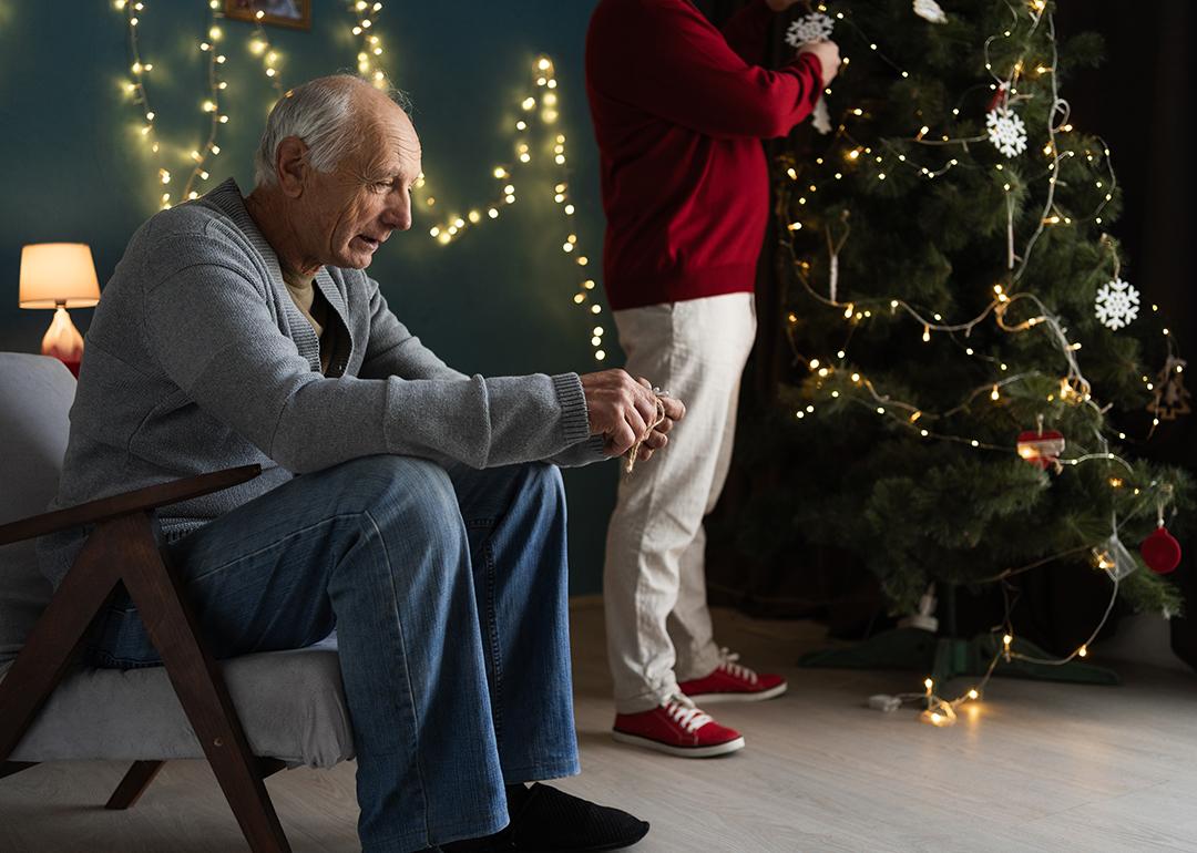 A stressed elderly man sitting by their Christmas tree as his partner decorates it.