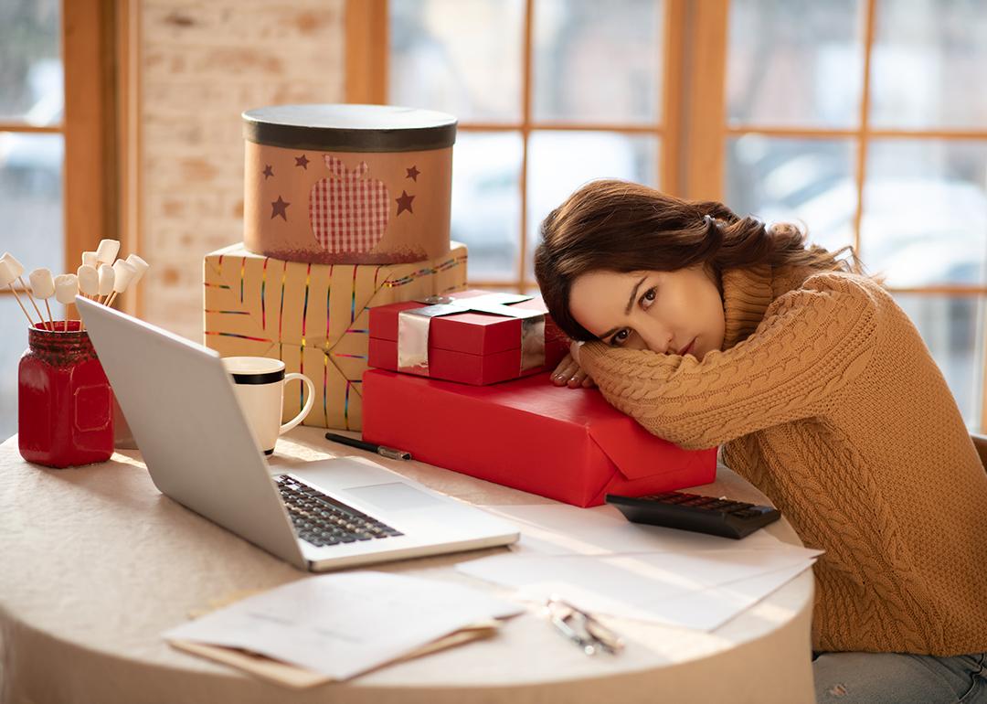 A young woman resting her head on a Christmas gift box alongside other presents in front of her laptop.