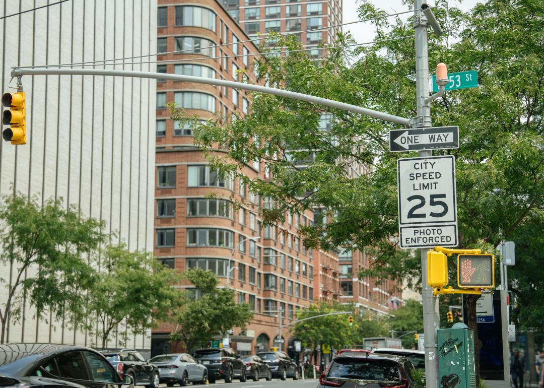A City Speed Limit 25 sign with a photo enforcement notice stands near a traffic light on a busy urban corner. Modern buildings, cars, and a street sign for 53rd Street are visible in the background.