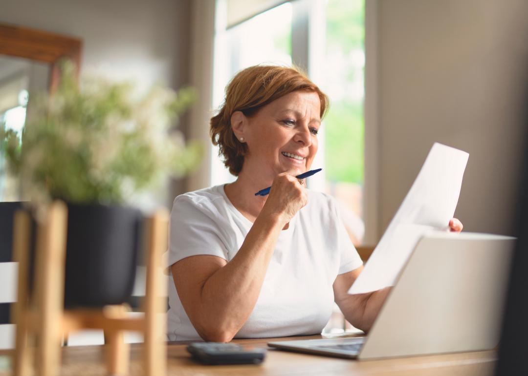 Middle-aged woman sitting at a table looking over paperwork with laptop open and pen in hand.