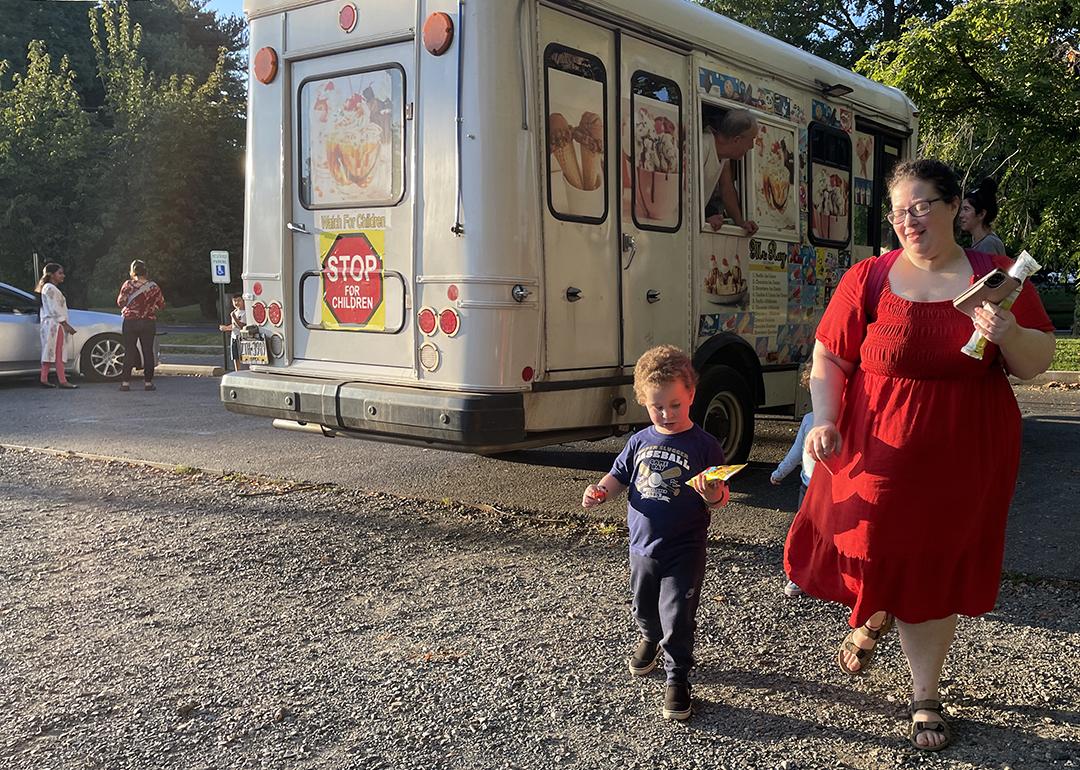 Kimberly Halevy wearing a crimson red midi dress with mid-length sleeves walks with her son Joshua beside her, enjoy treats from an ice cream truck visible behind them, after a recent session with a special instructor.