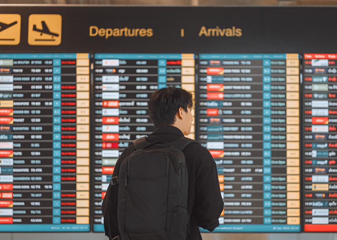 Rear view of a young man with a backpack looking at an airport's flight schedule board.