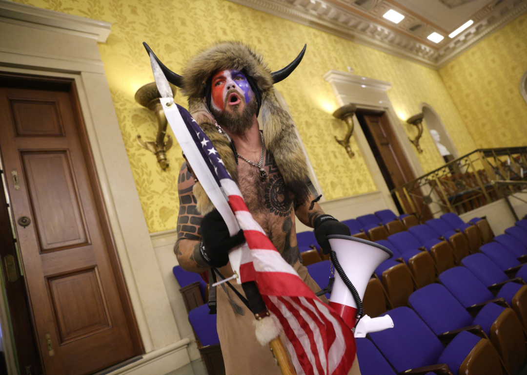 During the Capitol attack, Jacob Chansley, shirtless and wearing a horned helmet and a fur hat with his face painted in red, white, and blue, yells while holding an American flag with a spear attached top the end and a megaphone.