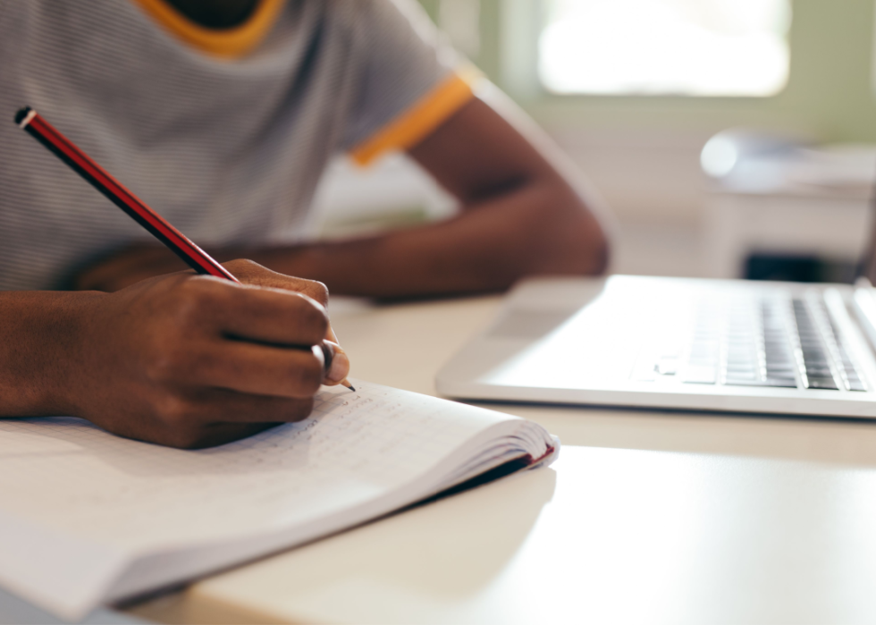 Student writing in notebook with laptop on desk
