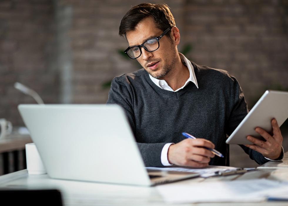 Man holding pen and tablet while looking at laptop.