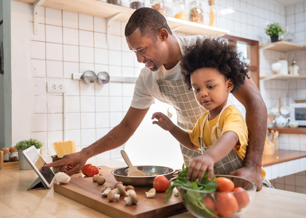 Man with son preparing meal with tomatoes, onions, and garlic, reading a recipe off a tablet..
