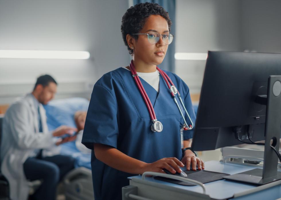 A medical professional works at a computer in a medical office with a doctor next to a patient in a hospital bed behind her..