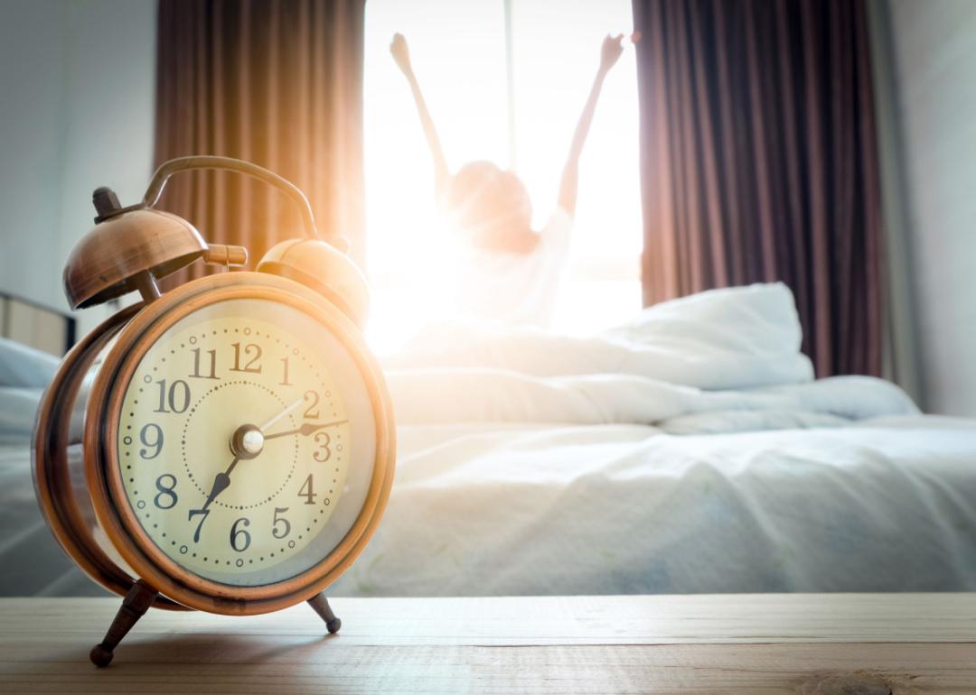 An alarm clock in the foreground and a waking teenager in the background.