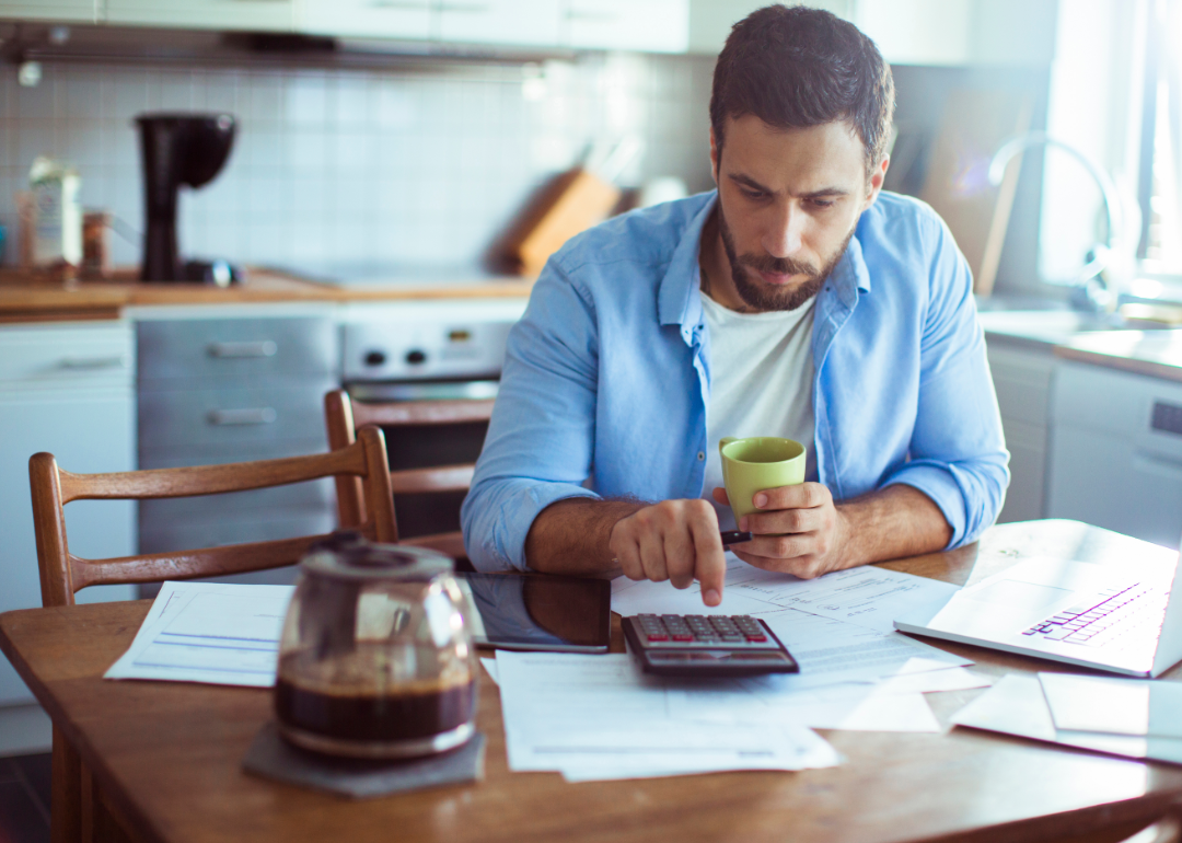 A man sits at the kitchen table with coffee, a calculator and a laptop.