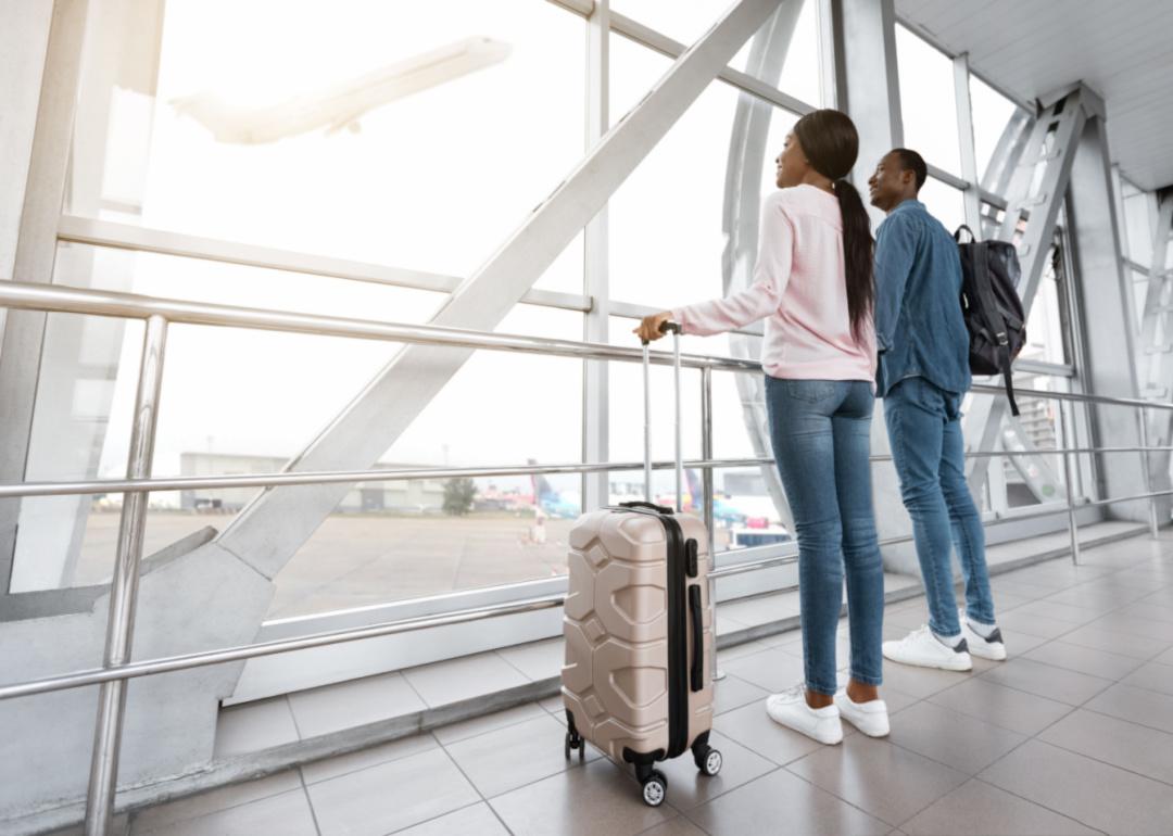 Millennial couple standing together near window In airport, looking at flying plane.