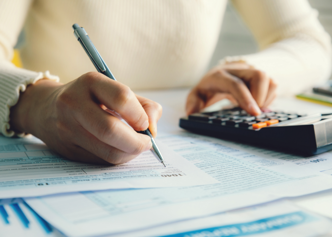 Two hands writing in a tax form and operating a calculator.