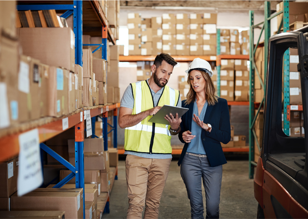Two workers wearing hard hats talk over a clipboard in a warehouse.