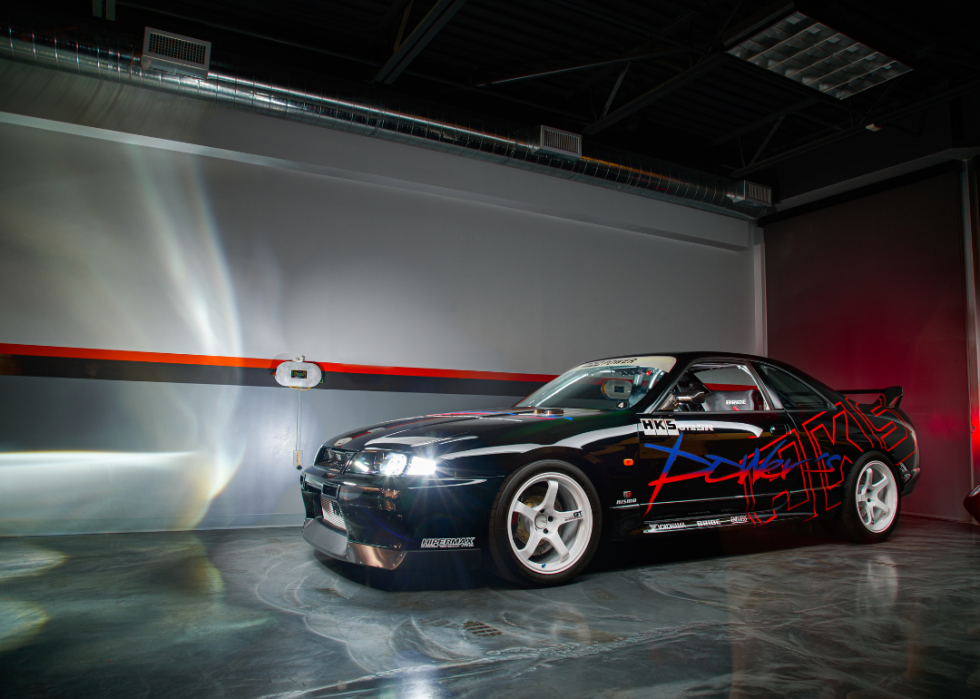 A modified black Nissan Skyline with its lights on in a parking area