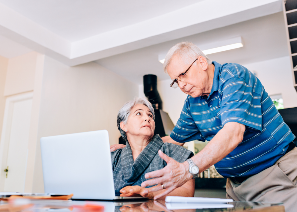 A worried senior couple at the kitchen table look at laptop.