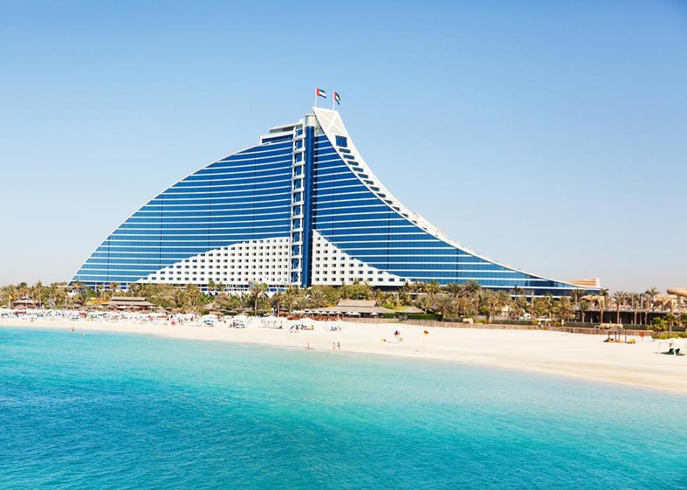 Jumeirah Beach hotel in Dubai with the pure white sand beach and crystal clear water in front of it surrounded by green palm trees.