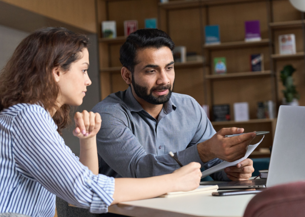 Two employees sit at a desk talking as they look at a laptop.