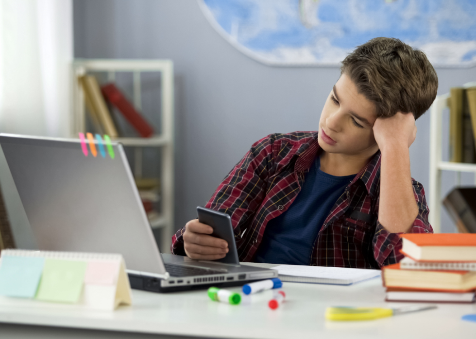 A young boy appears disinterested as he sits at his desk, in front of the computer, engrossed in his cell phone.