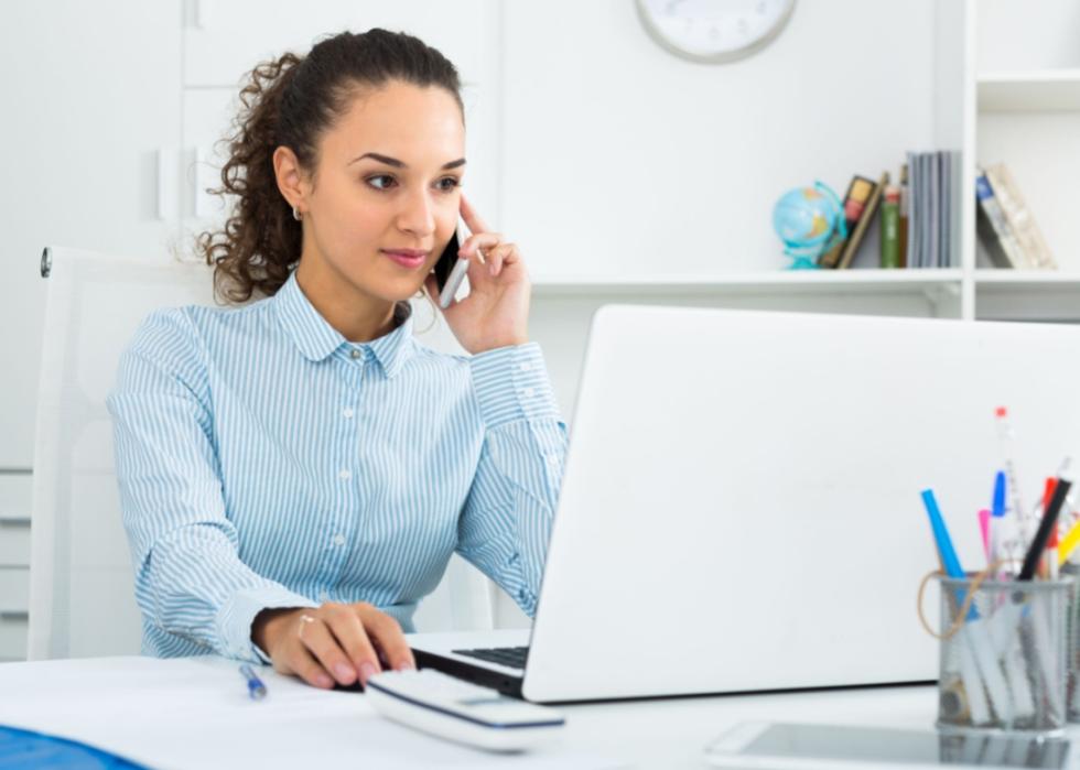 A young woman in a business attire sitting at the desk holding a phone close to her ear.