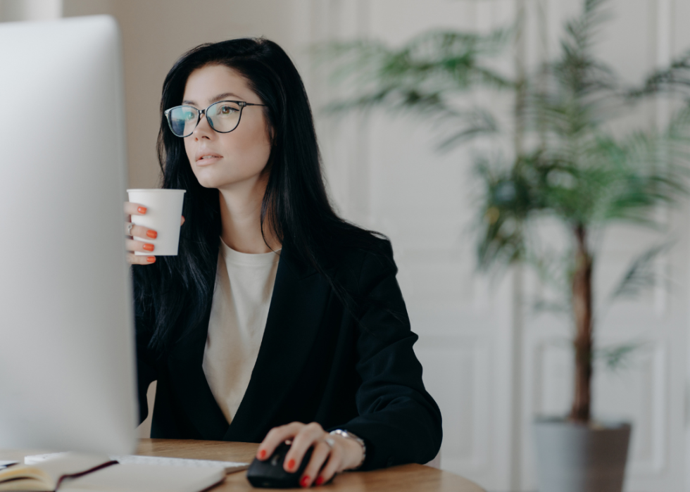 A young woman sitting at a desk looking at the computer while holding a cup.