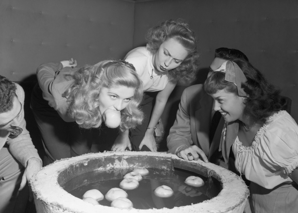 A black and white photo of a group of people dunking for apples. A woman in the center has an apple clenched firmly between her teeth.