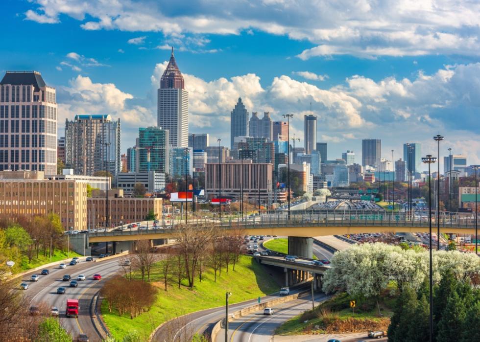 City skyline dominated by modern, gklass skyscrapers of varying heights. A multi-lane highway with heavy traffic with cars and trucks in the foreground.