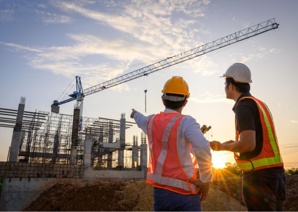 Two men wearing ornage vests and helmets at the construction site with tall metal rods and a tall crane.
