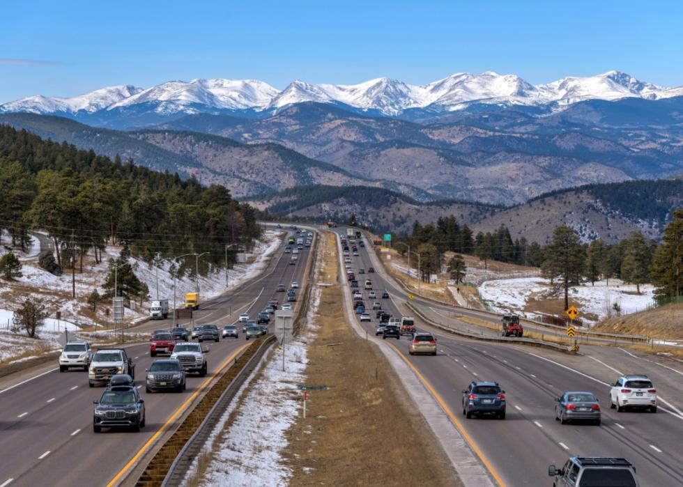A highway with multiple lanes cuts through landscape with tall snowy mountaiuns in the distance.
