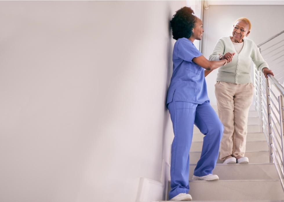 An elderly woman walking down the stairs with the help of a careworker in blue scrubs. 