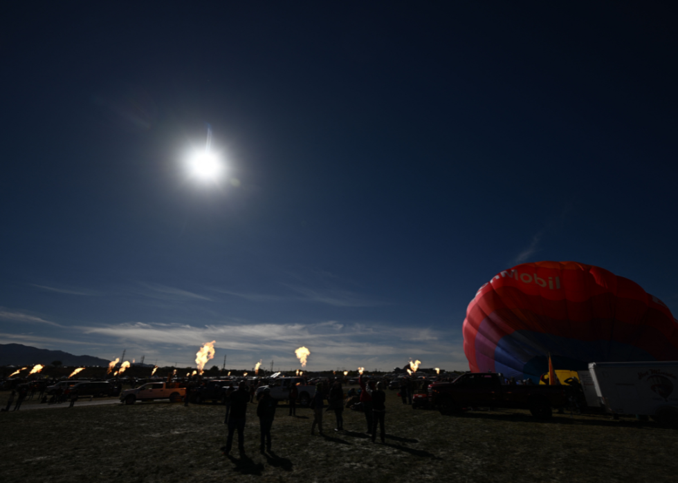 A crowd of people gathered in the field standing and looking up at the dark sky with a few fire pits burning in the background. 