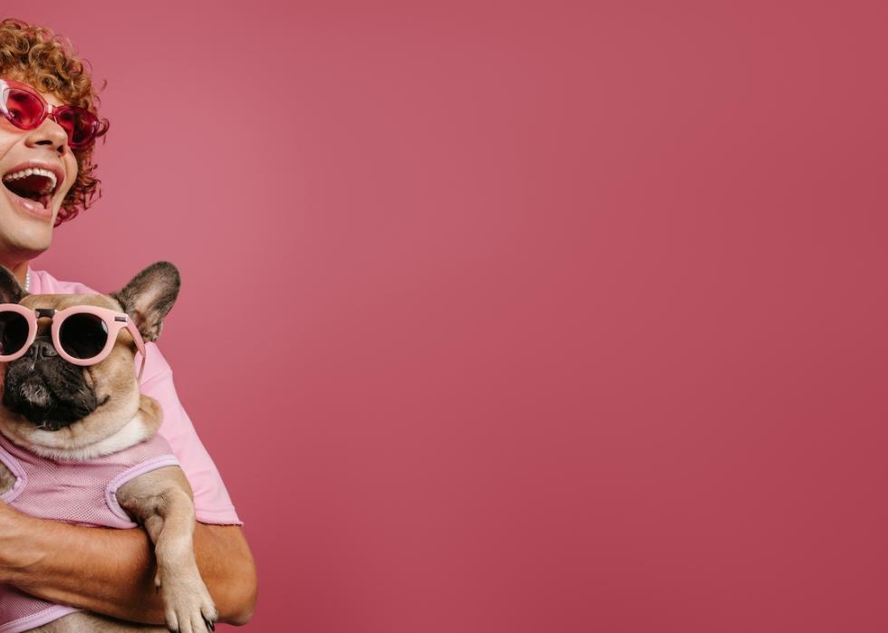 Joyful young person with curly hair holds a French bulldog, as both wear pink sunglasses on a pink background.