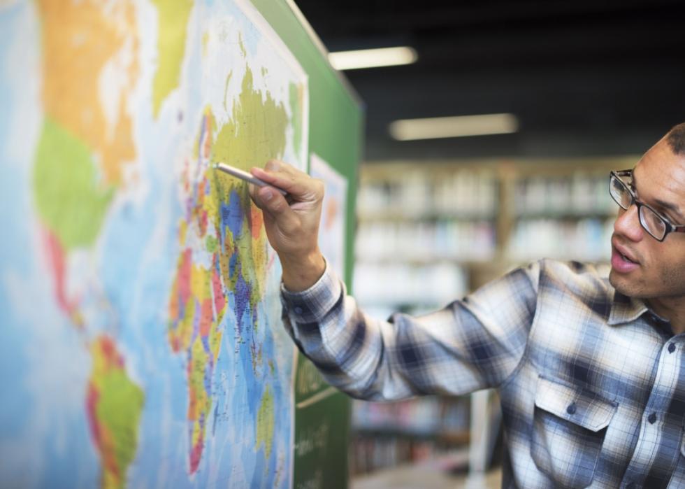A young African American teacher with glasses is pointing at the world map in front of him.